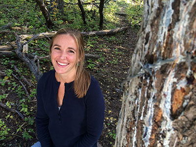 Smiling woman sitting outdoors near a tree, surrounded by greenery, showcasing a joyful moment in nature.